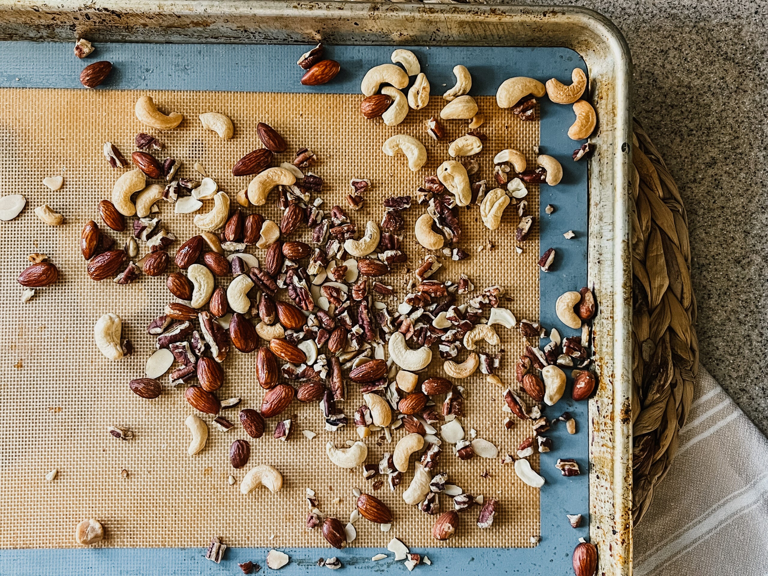 A close-up of a baking sheet with a mixture of roasted cashews, walnuts, and almonds. The sheet sits on a rattan placemat and tea towel.