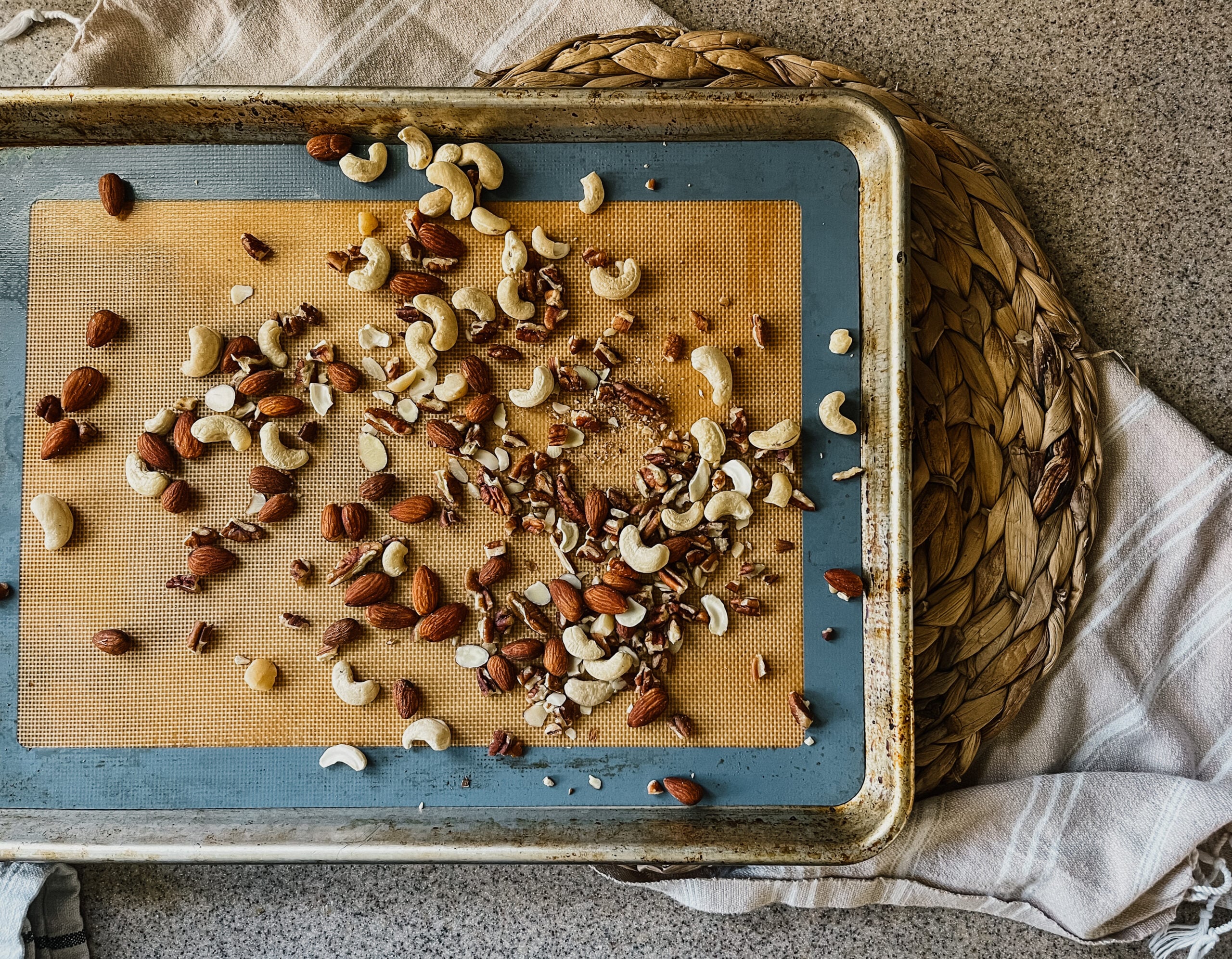 A baking sheet with a mixture of roasted cashews, walnuts, and almonds. The sheet sits on a rattan placemat and tea towel.
