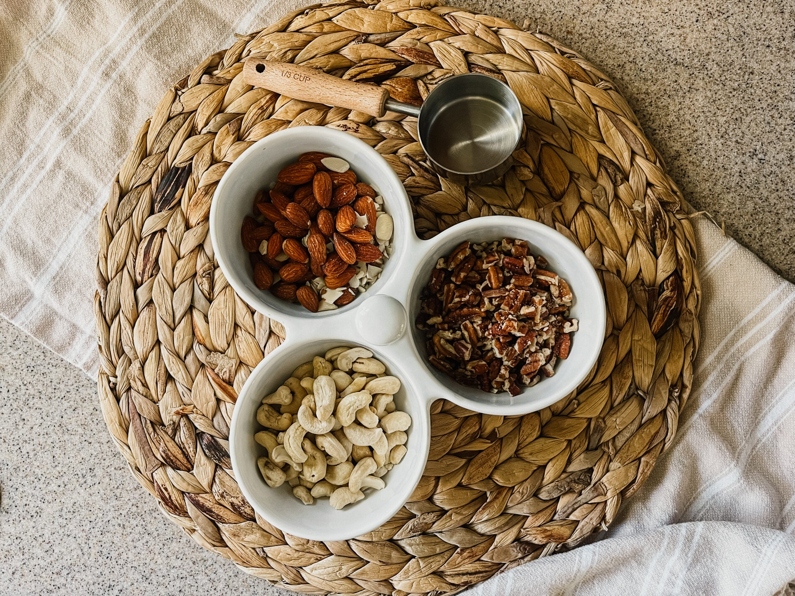 Three bowls with almonds, walnuts, and cashews on top of a rattan placemat. A measuring cup sits towards the top of the image.