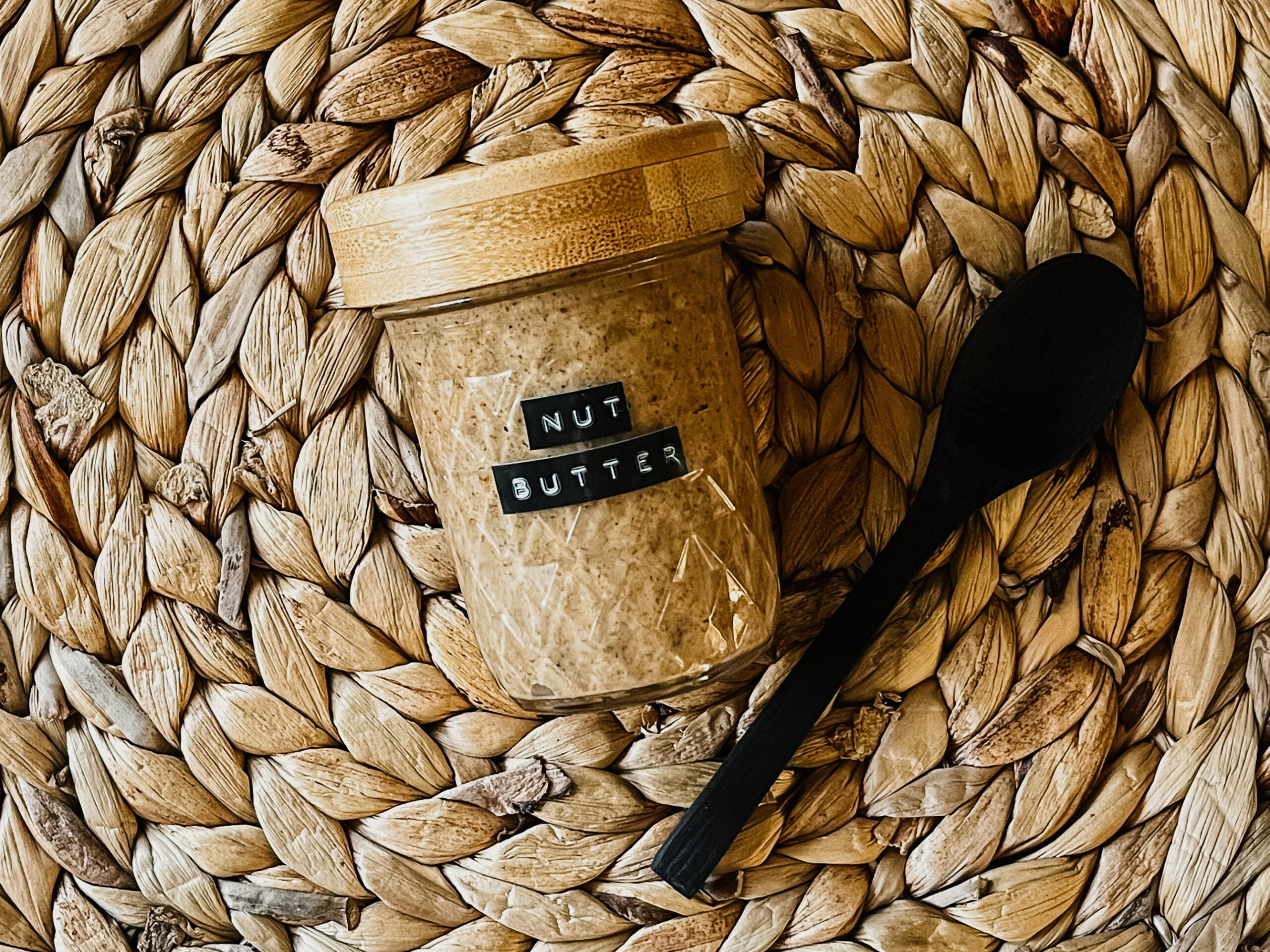 A jar with a wood top and label that states “Nut Butter.” A wood spoon sits to the right and both are on top of a rattan placemat.