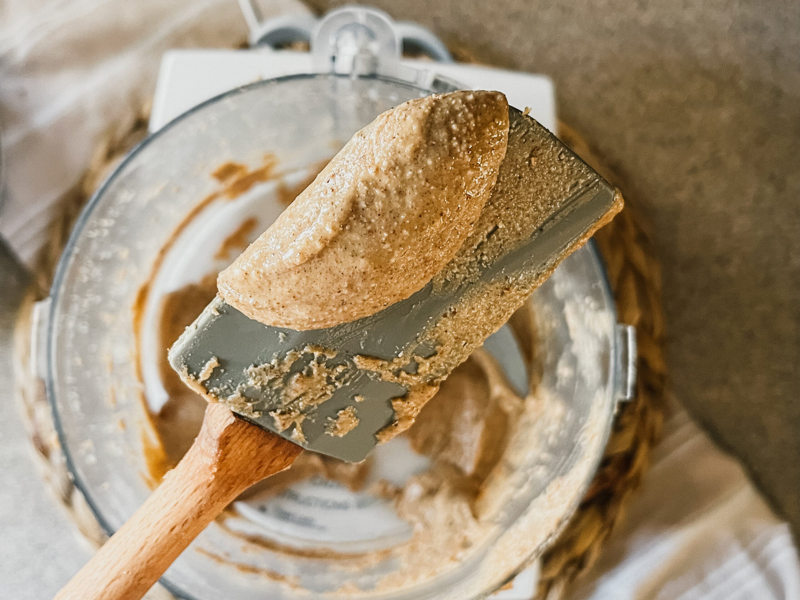 A spatula with a scoop of completed, creamy nut butter. A food processor with the remaining nut butter is in the background.