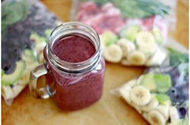 A berry smoothie in a mason jar cup on a table surrounded by prepped smoothie freezer bags.