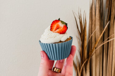 A hand holding a frosted almond flour cupcake. A strawberry slice sits on top of the cupcake and a palm frond is in the background.