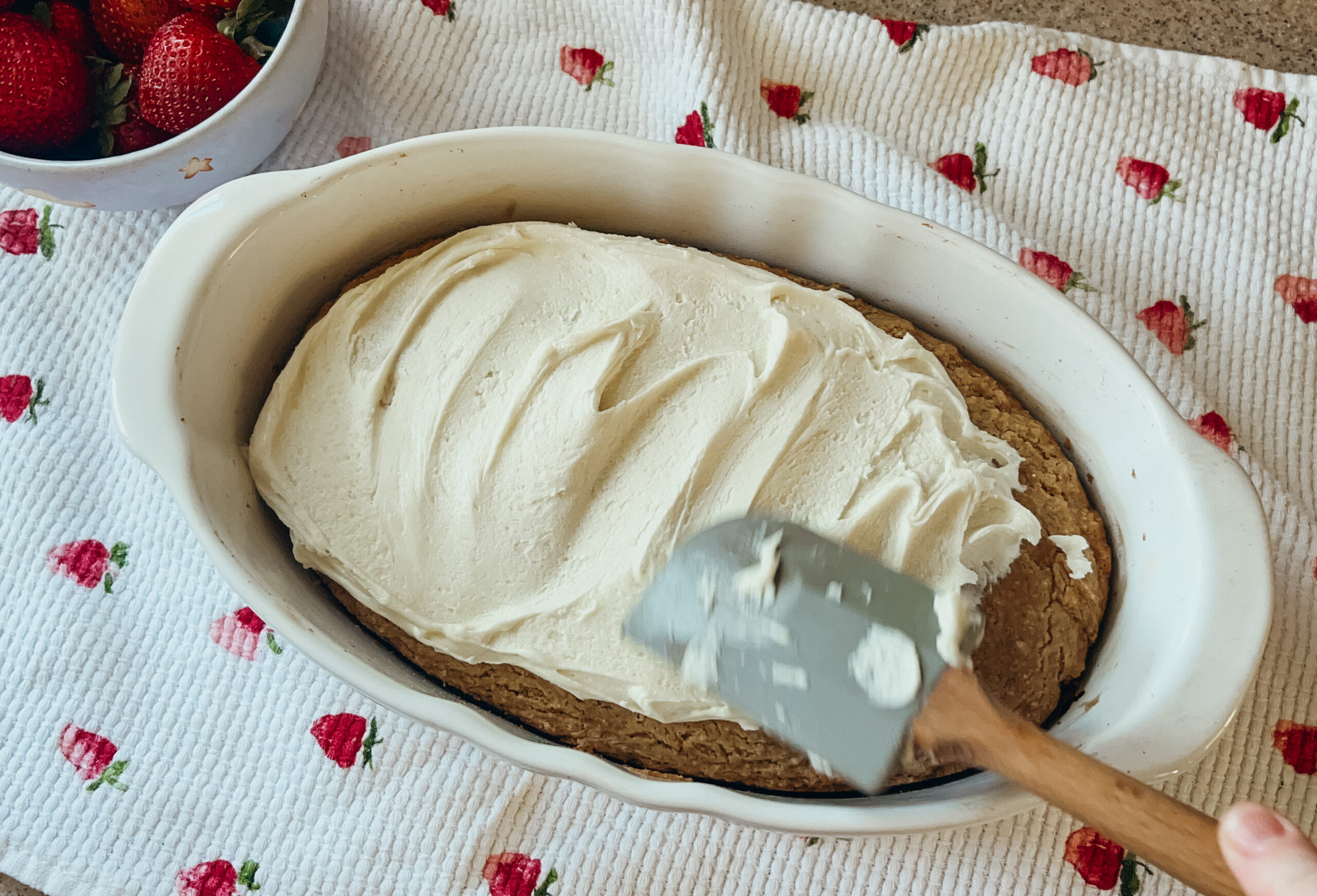 A strawberry shortcake being frosted with a blue spatula. A strawberry printed tea towel sits underneath.