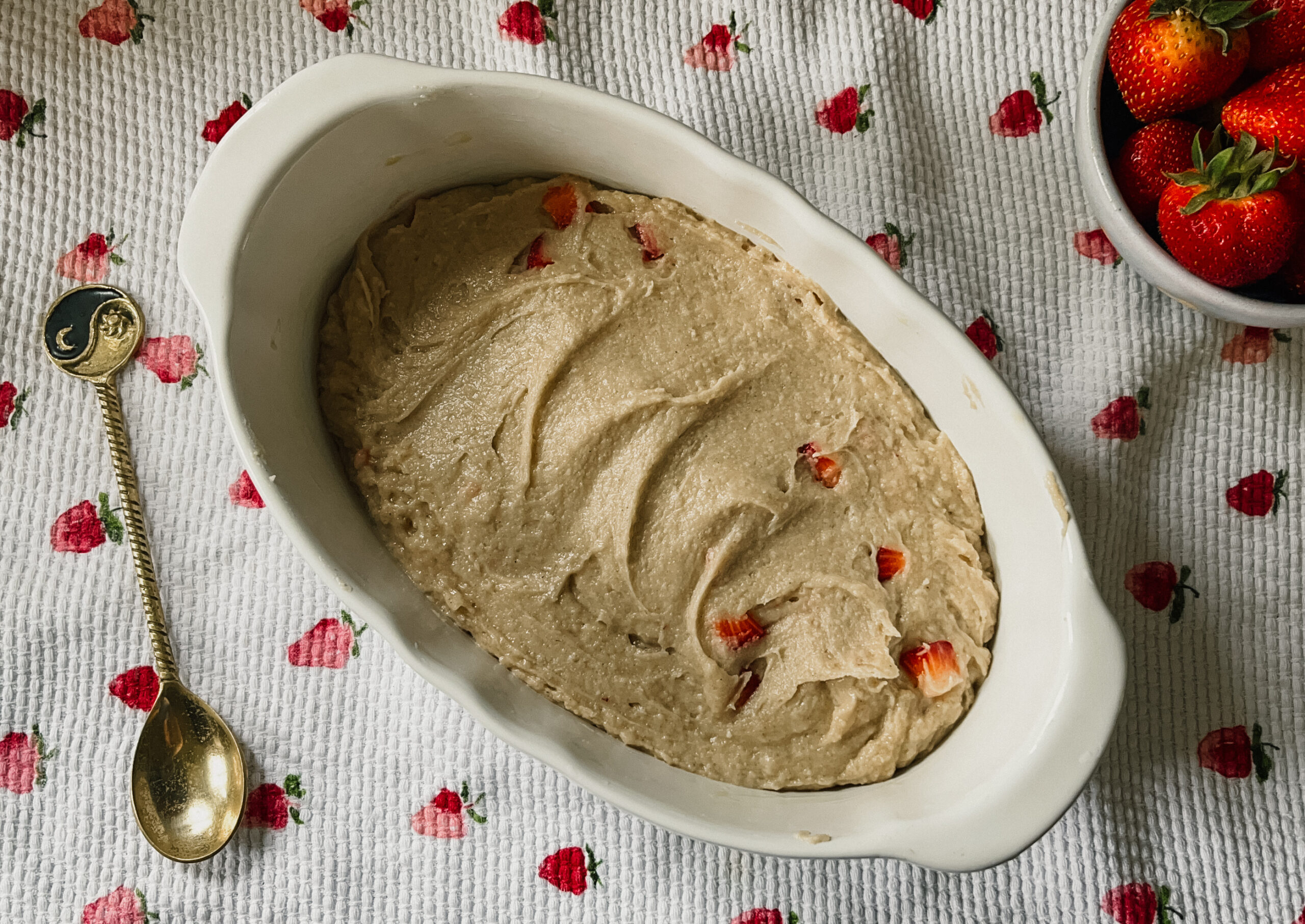 An oval dish filled with strawberry shortcake batter. A strawberry printed tea towel sits underneath.