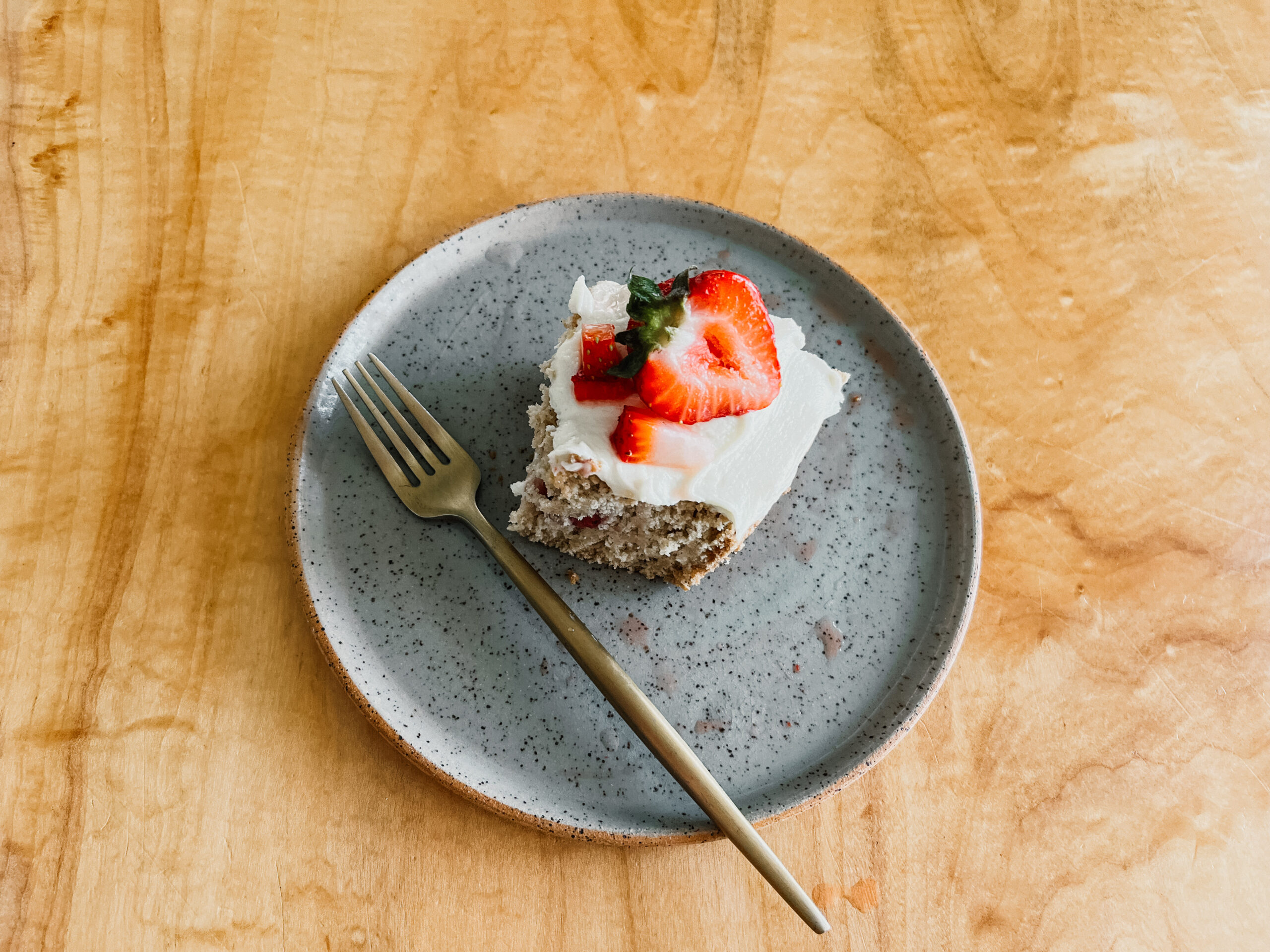 A slice of gluten-free strawberry shortcake on a plate with a gold fork. Sliced and chopped strawberries sit on top nestled in the frosting. 