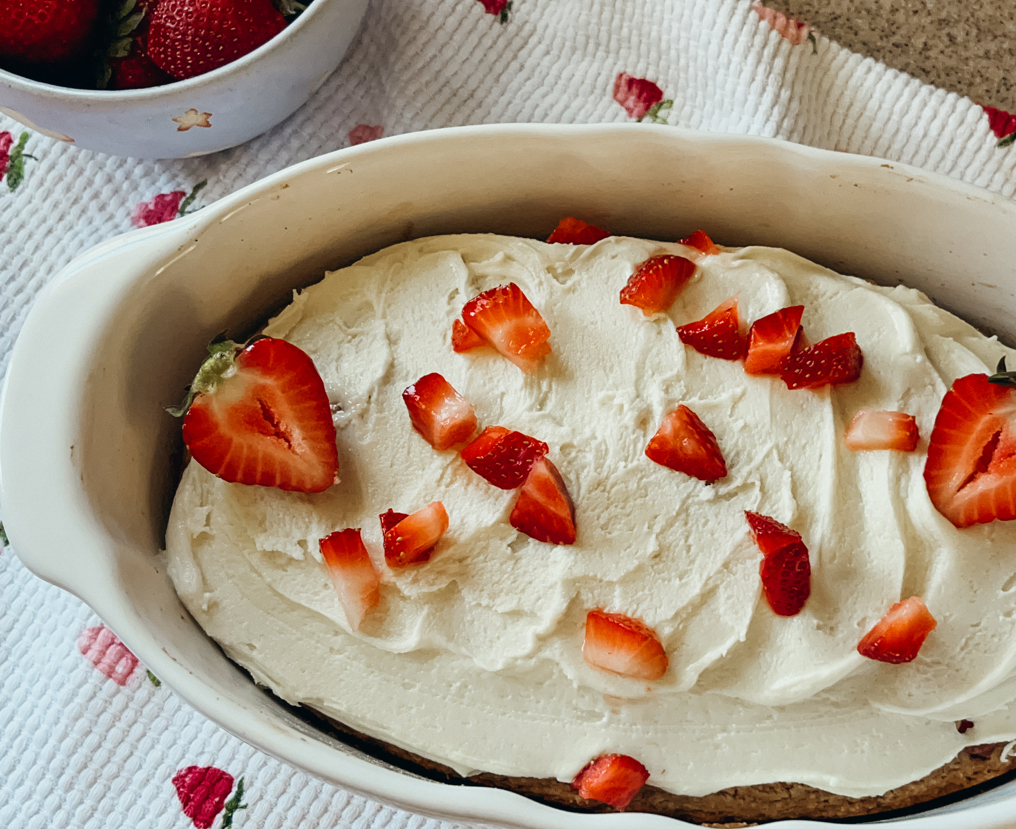A close up of a freshly frosted strawberry shortcake. Chopped and sliced strawberries are sprinkled on top of the frosting. A strawberry printed tea towel sits underneath.