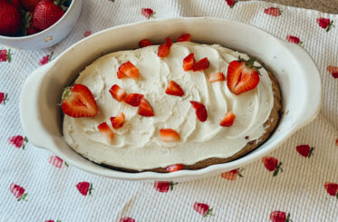 An oval shaped dish of a freshly frosted strawberry shortcake. Chopped and sliced strawberries are sprinkled on top of the frosting. A strawberry printed tea towel sits underneath.