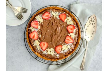 A chocolate pie topped with sliced strawberries, almonds, and coconut next to a serving spoon and plate with forks.