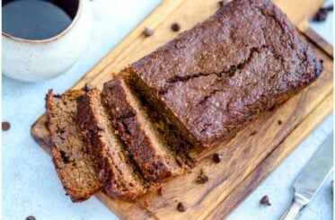 A chocolate zucchini bread loaf on a wooden cutting board with chocolate chips sprinkled around it beside a cup of coffee.