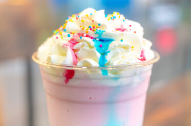 Pink milkshake topped with whipped cream, blue and red syrup, and colorful sprinkles.