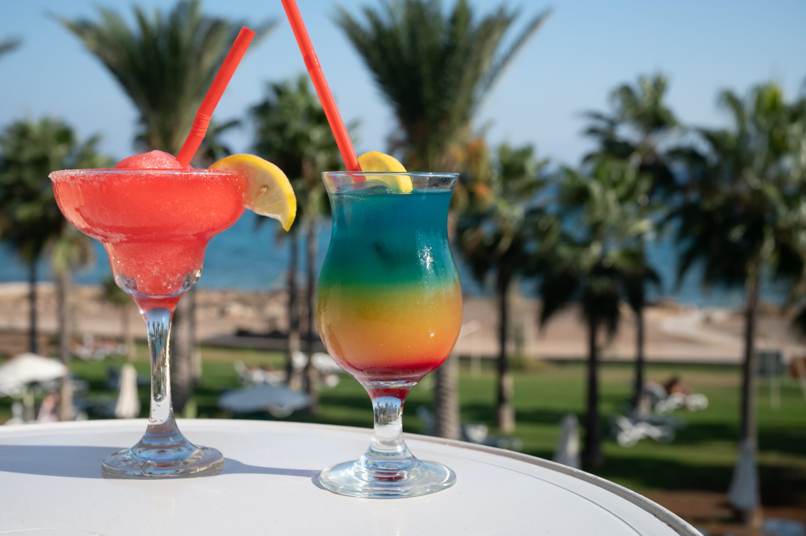 Colourful,Cold,Strawberry,Daiquiri,And,Rainbow,Paradise,Cocktails,Drinks,Served Rainbow and strawberry daiquiris against a beach backdrop.