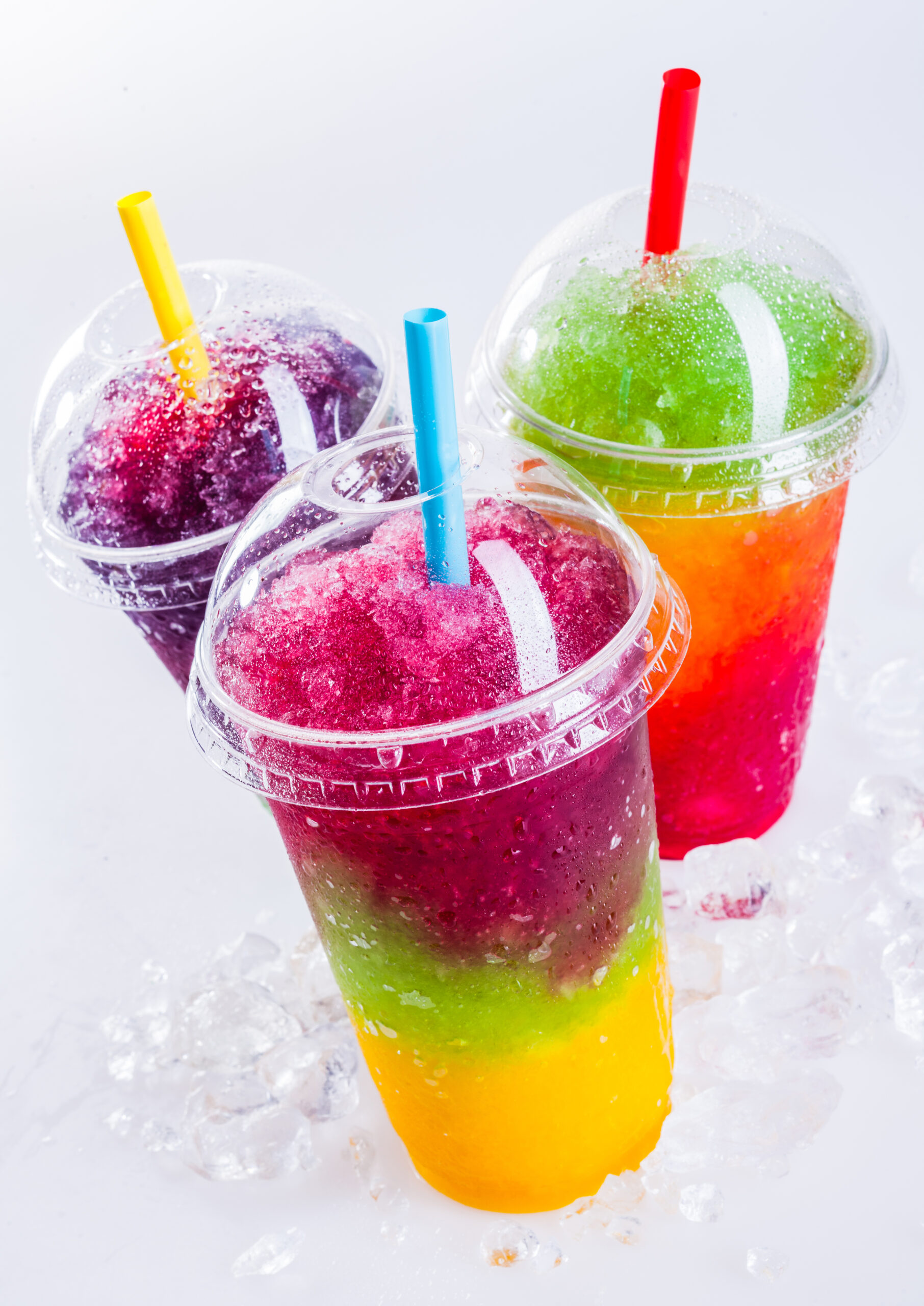 Rainbow,Layered,Slush Three plastic cups containing rainbow layered sluch on a white ice-covered surface.