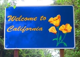 Road sign says "Welcome to California" with yellow flowers and a blue background to the sign