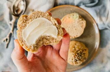 A hand holding a slice of roll slathered in butter with a bite taken. A plate with the remaining rolls sits behind the hand on a blue tea towel. A measuring spoon is in the top left.