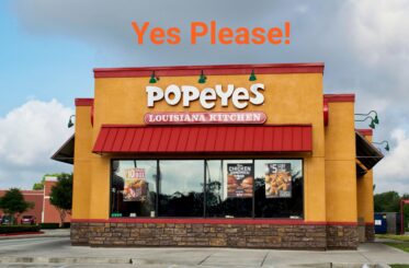 Popeyes restaurant and logo with "Yes Please!" written above in orange letters. Clouds and sky are behind.