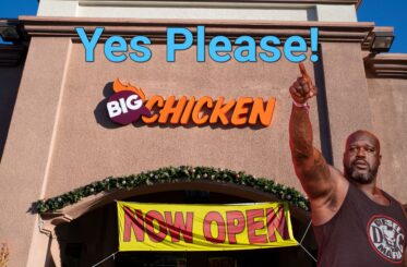 Shaq pointing up at "Yes Please" in from of a Big Chicken restaurant with a "Now Open" sign.