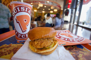 Chicken sandwich with Popeyes drink next to it and people in the background