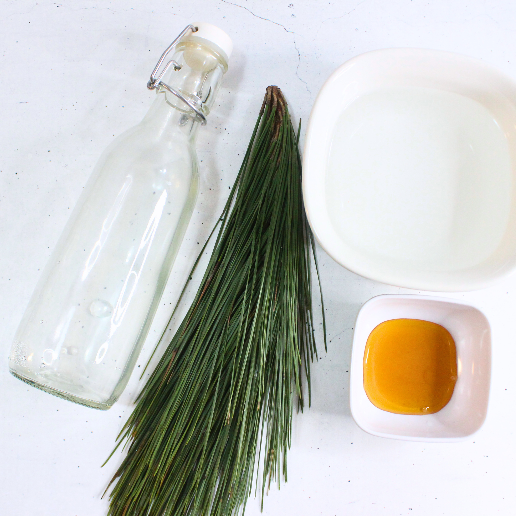 Pine needles, honey, water, and a glass bottle on a white countertop.