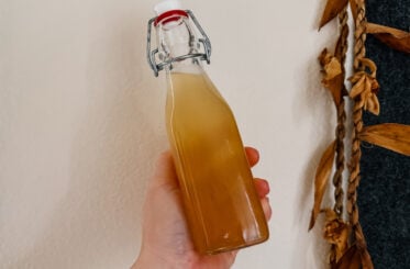 A hand holding a bottle of apple cider vinegar and honey. Dried flowers hang to the right side of the image.