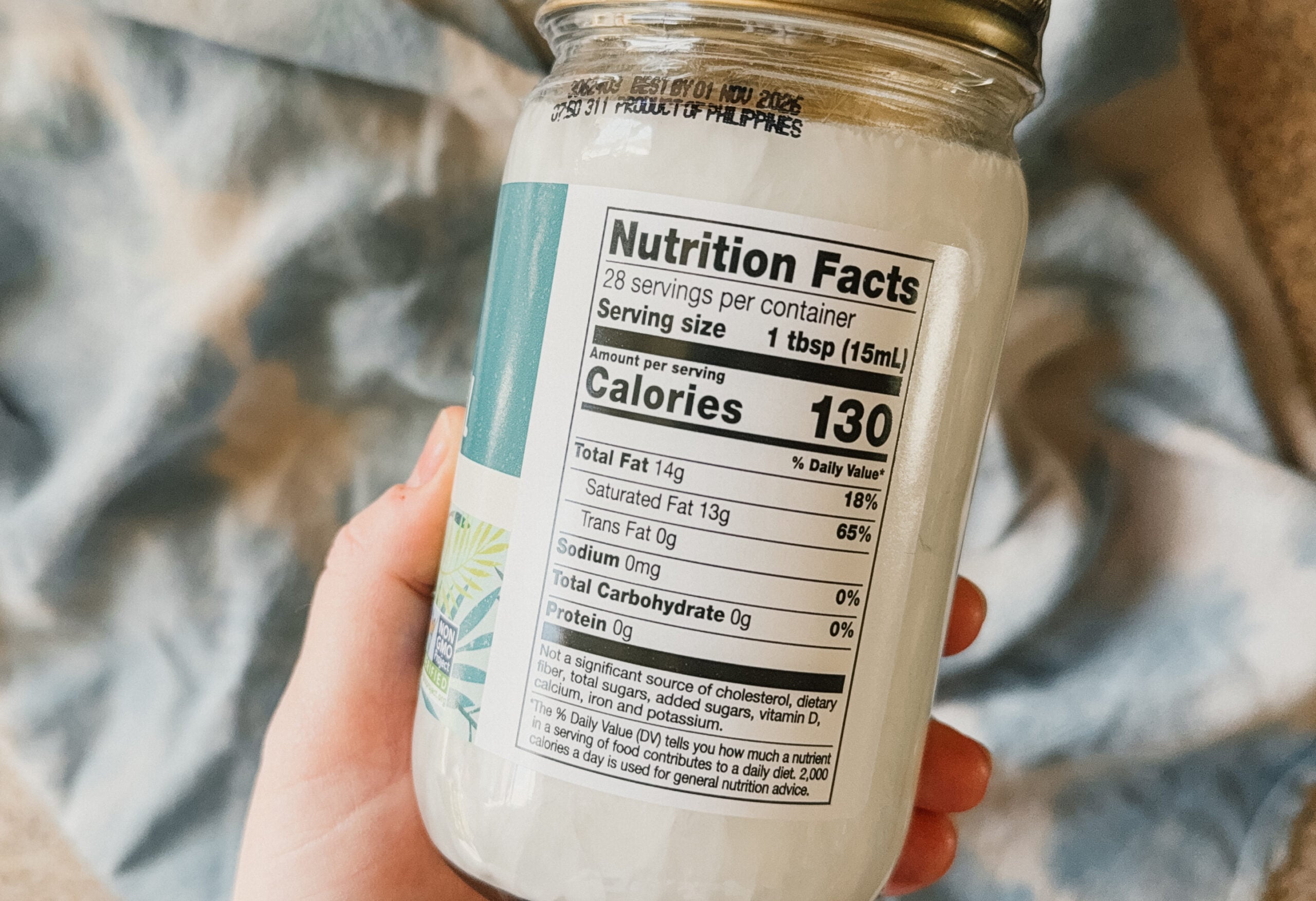 A hand holding a jar of coconut oil with the nutrition facts facing forward. A blue tea towel lays beauty.