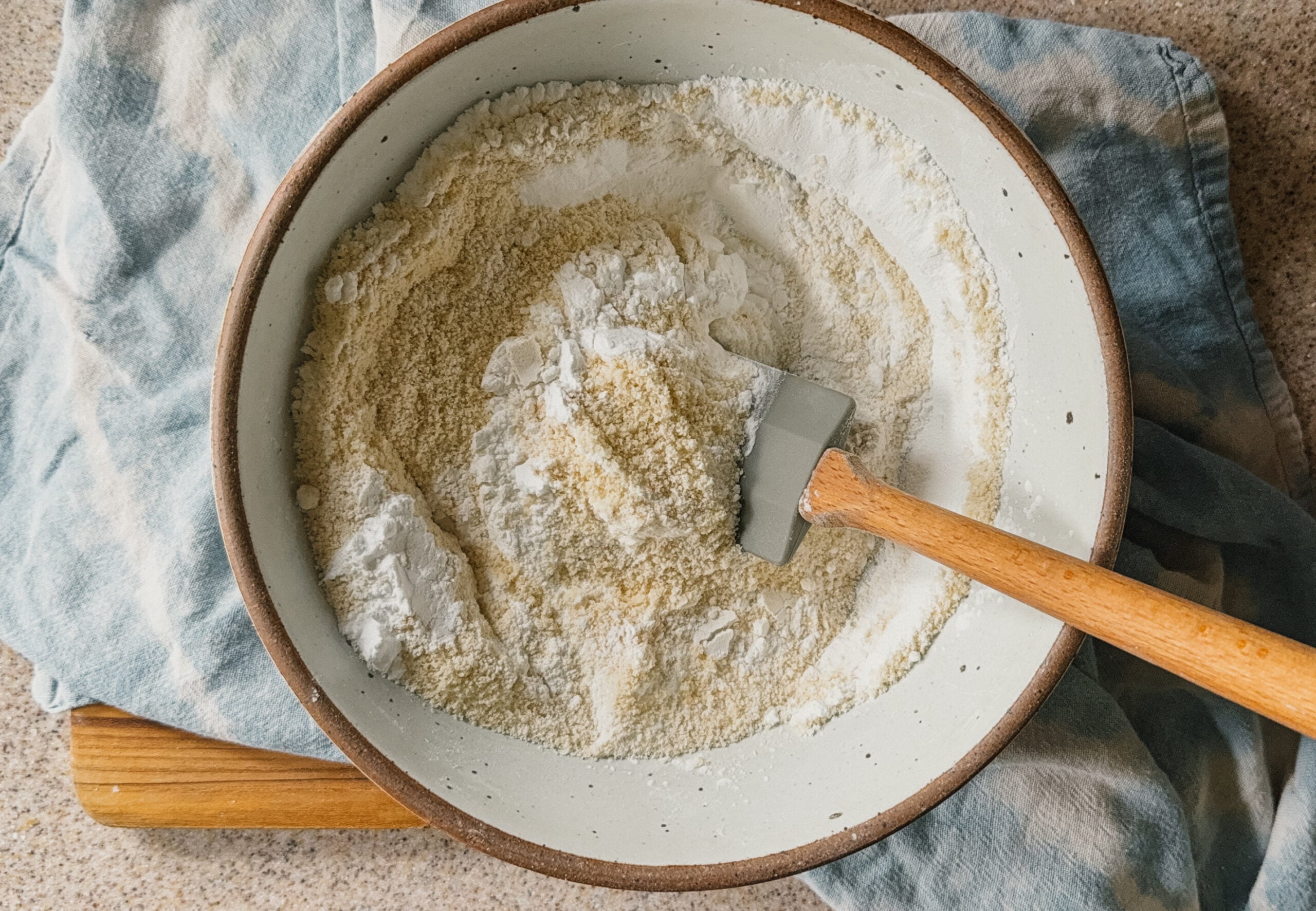 A bowl with ingredients being mixed. A spatula sticks out of the right side of the bowl and a blue tea towel is underneath it.
