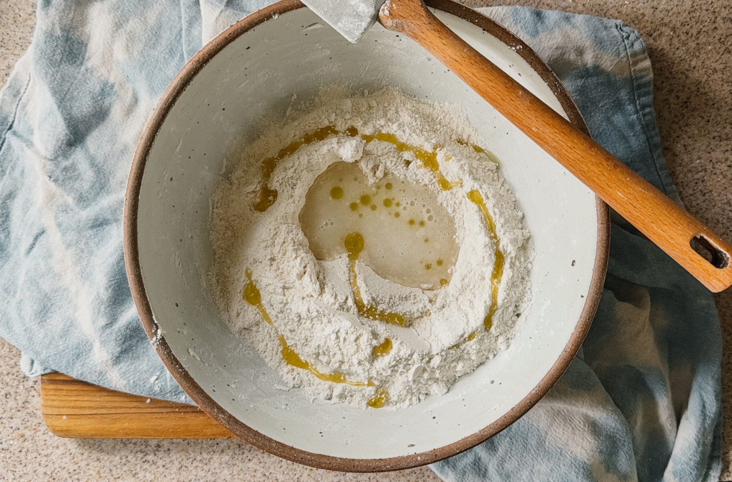 A bowl with ingredients about to be stirred. A spatula rests on the top right of the bowl and a blue tea towel is underneath it.