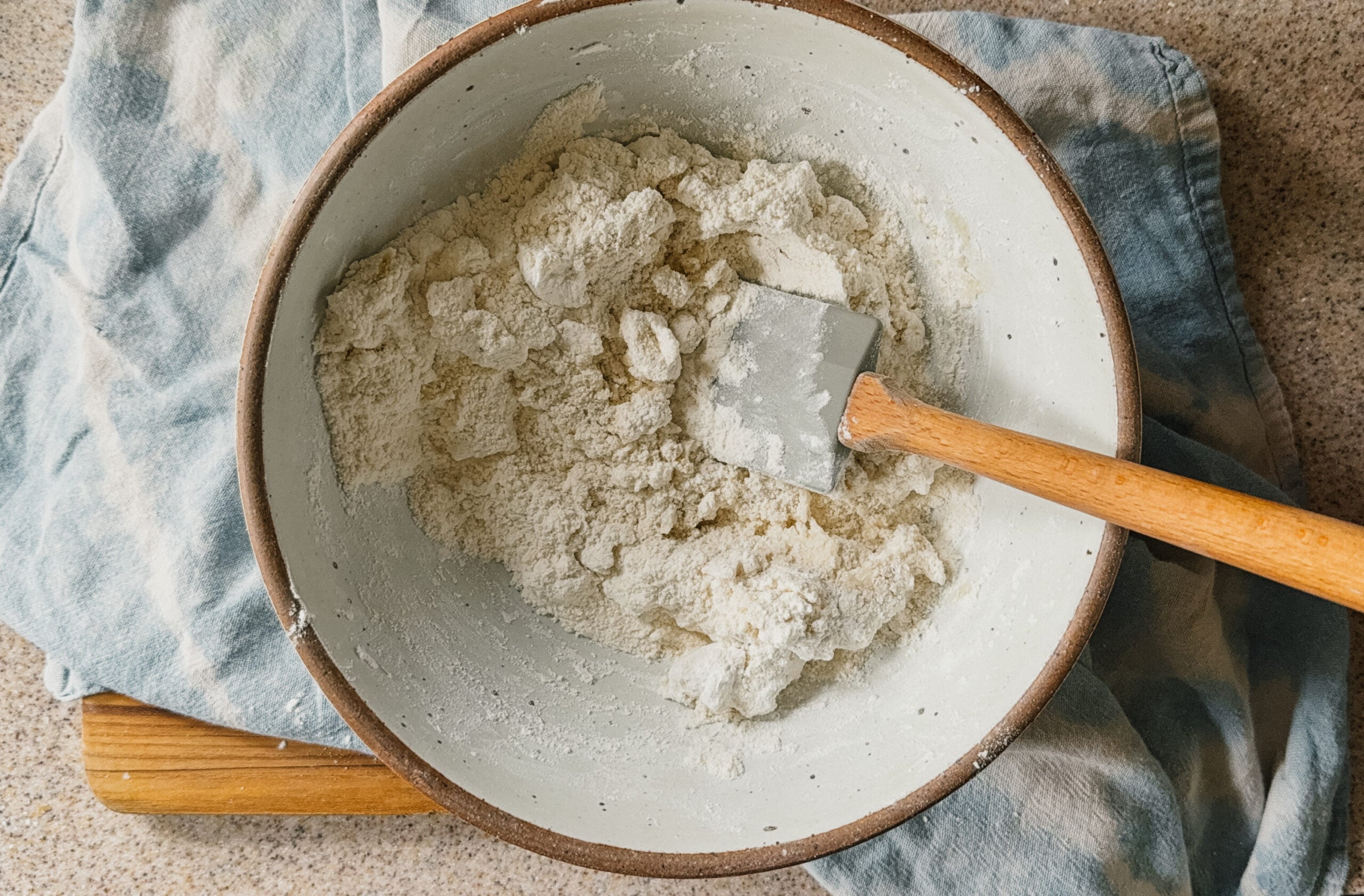 A bowl with ingredients being mixed. A spatula sticks out of the right side of the bowl and a blue tea towel is underneath it.