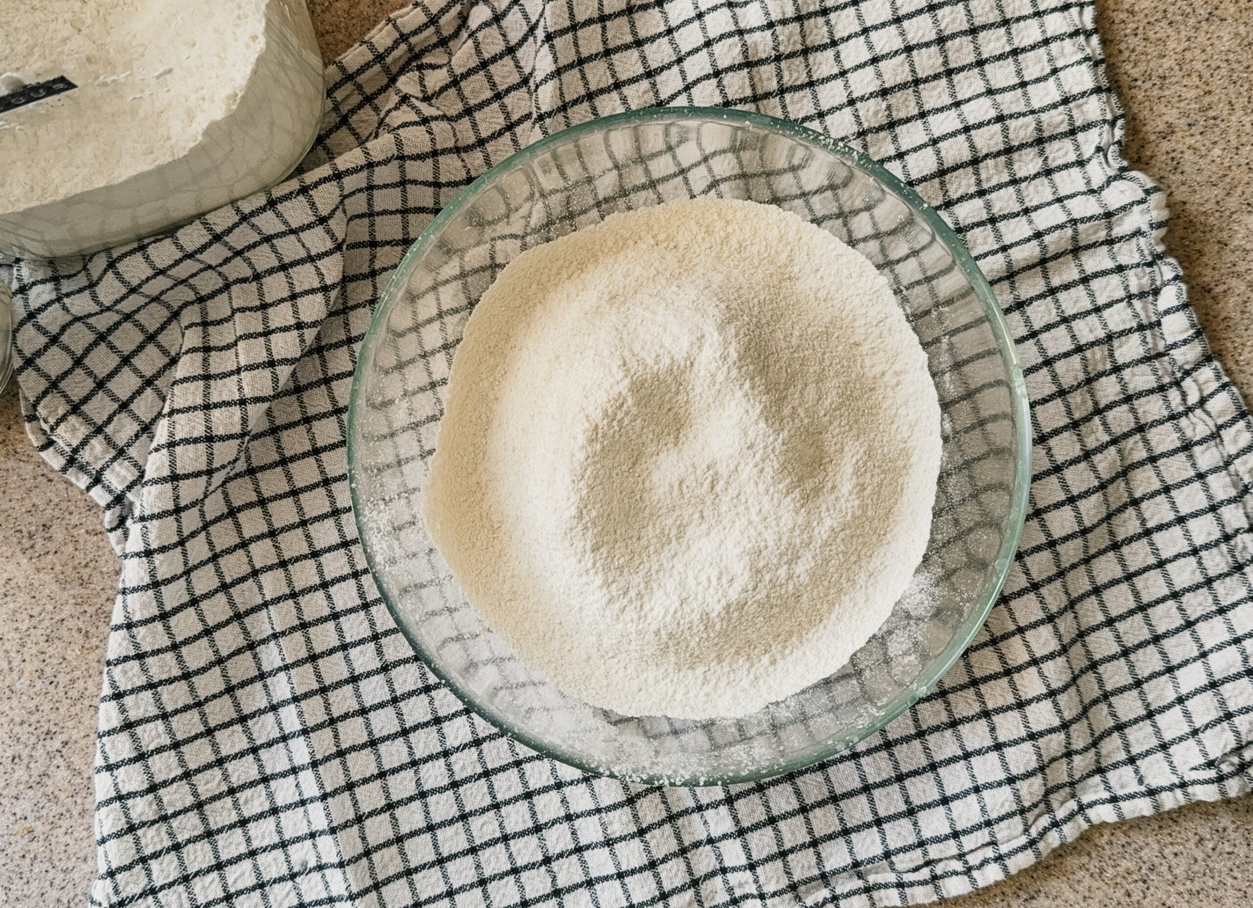A sifted flour mix in a glass bowl. A checkered tea towel sits underneath.