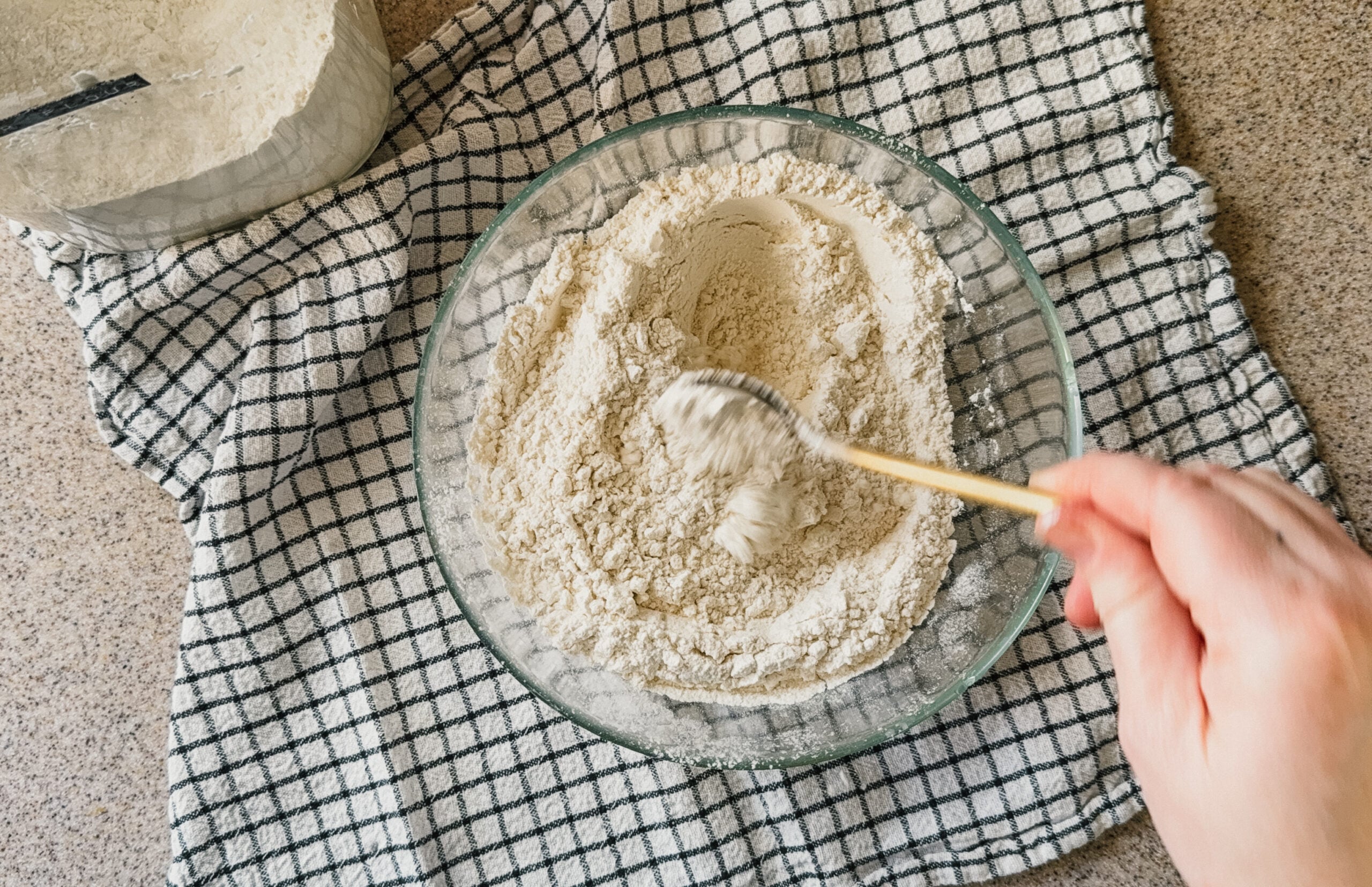 A hand mixing flour in a bowl with a gold spoon. A checkered tea towel sits underneath.