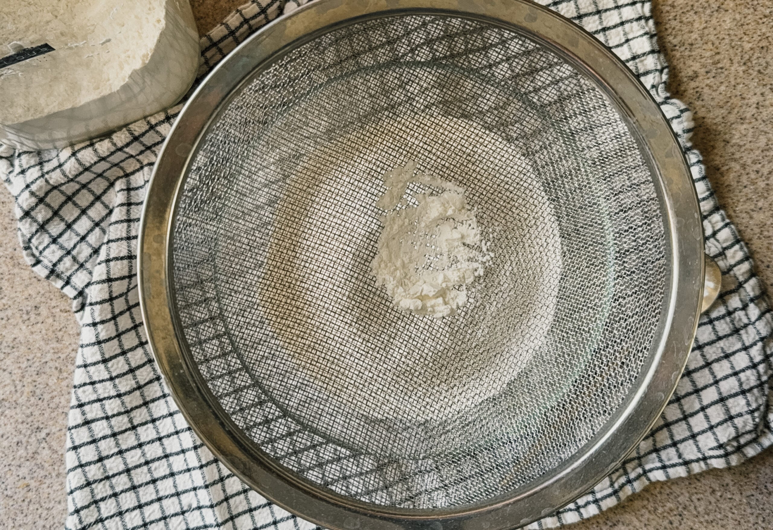 Cornstarch being sifted through a strainer into a bowl. A checkered tea towel sits underneath.