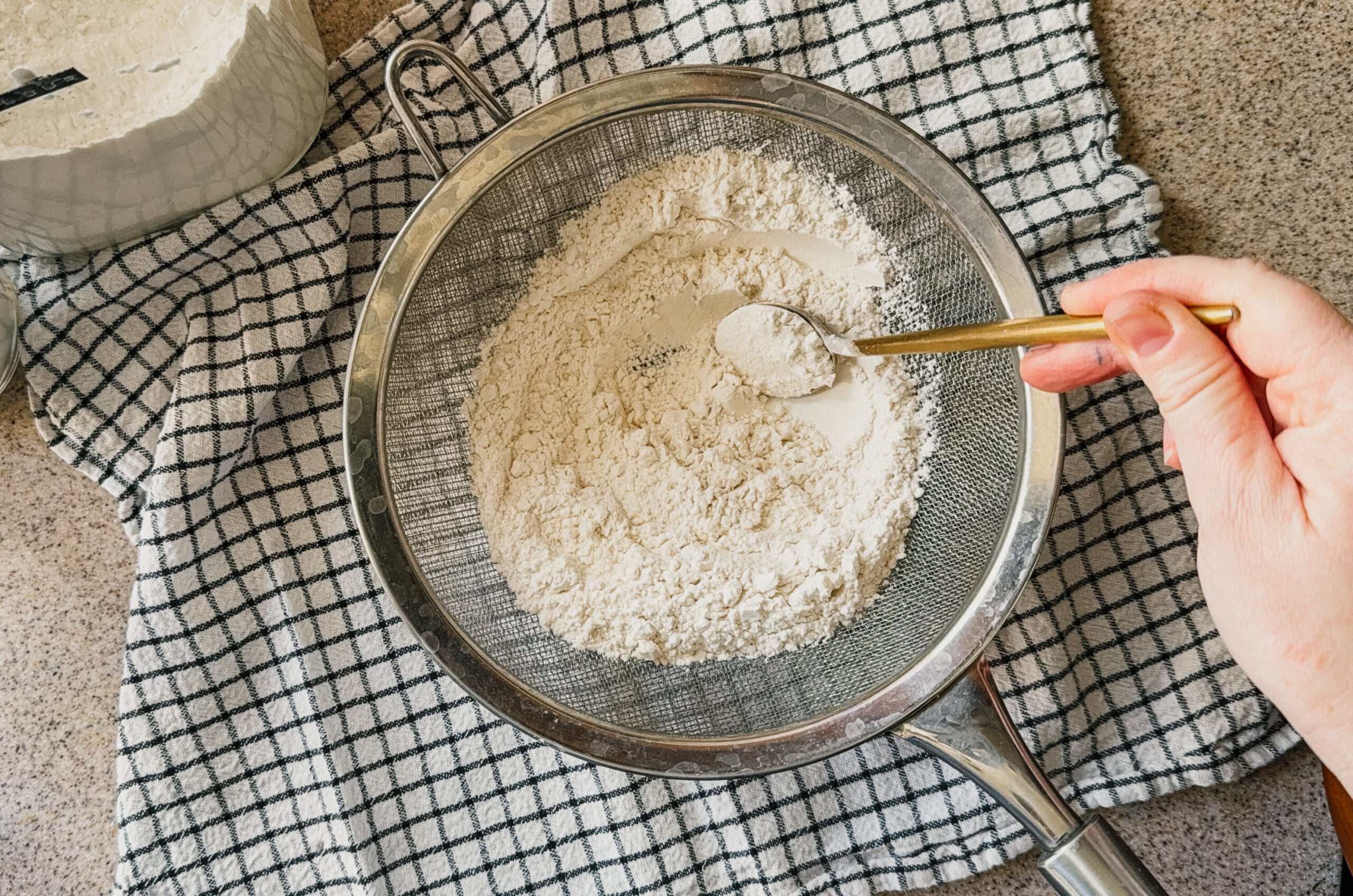 A hand sifting flour through a strainer with a gold spoon. A checkered tea towel sits underneath.