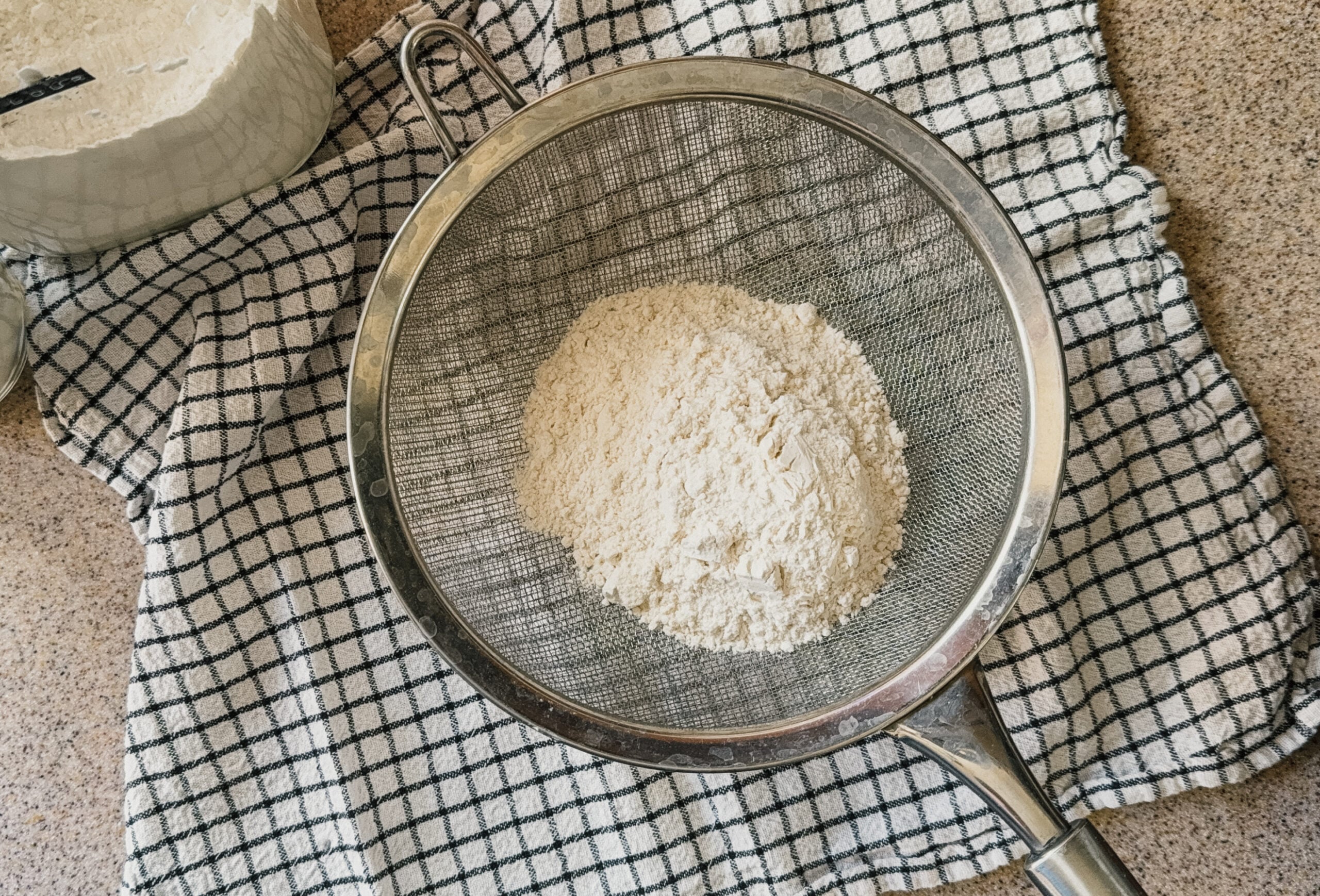 A strainer with a cup of flour about to be sifted into a bowl. A checkered tea towel sits underneath.