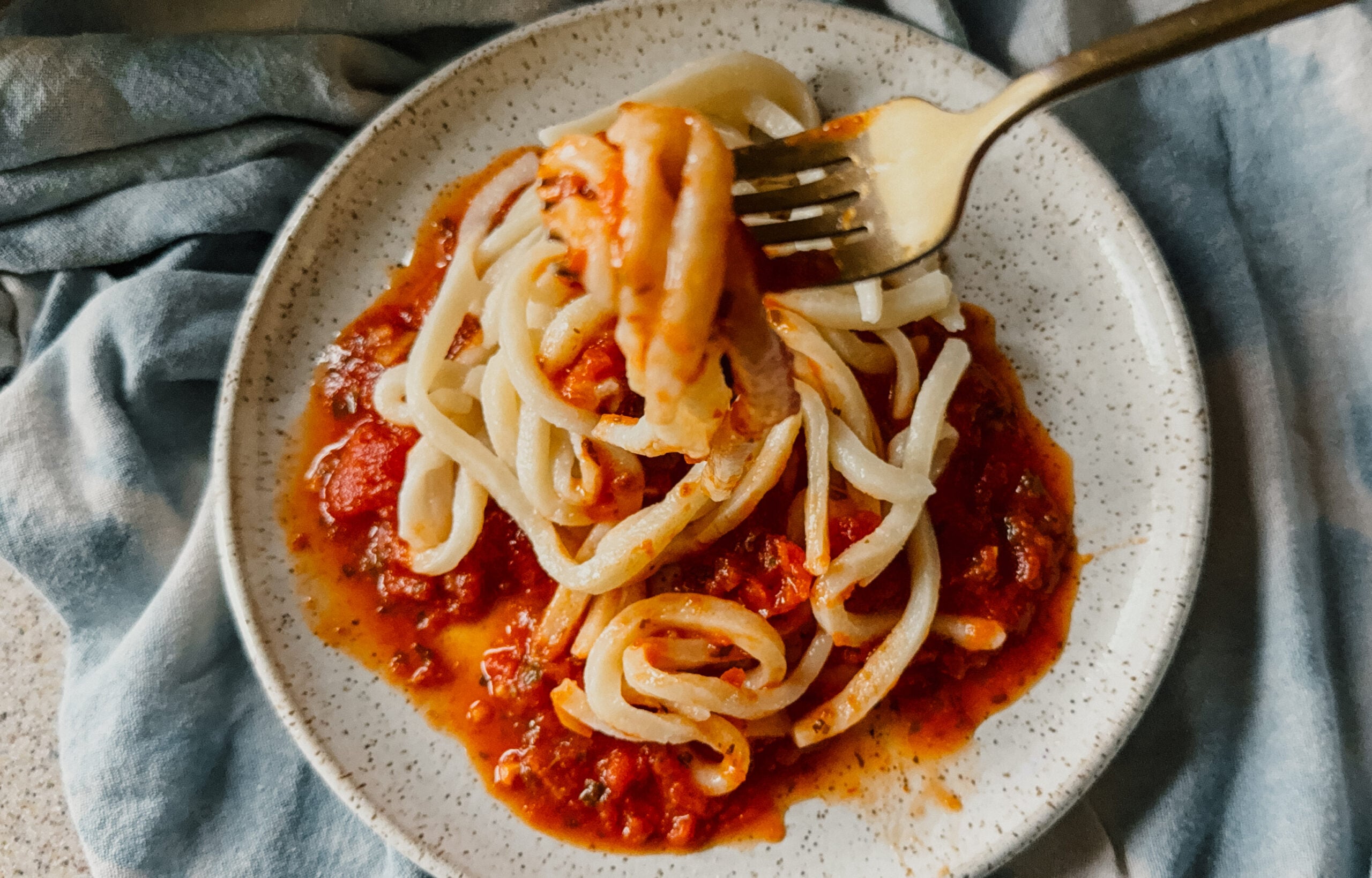 A plate of noodles with red sauce. A gold fork lifts some of the noodles from the plate.