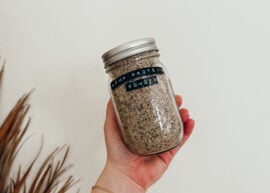 A hand holding a jar of hemp protein powder in front of a white wall. A palm frond is in the bottom left of the image.