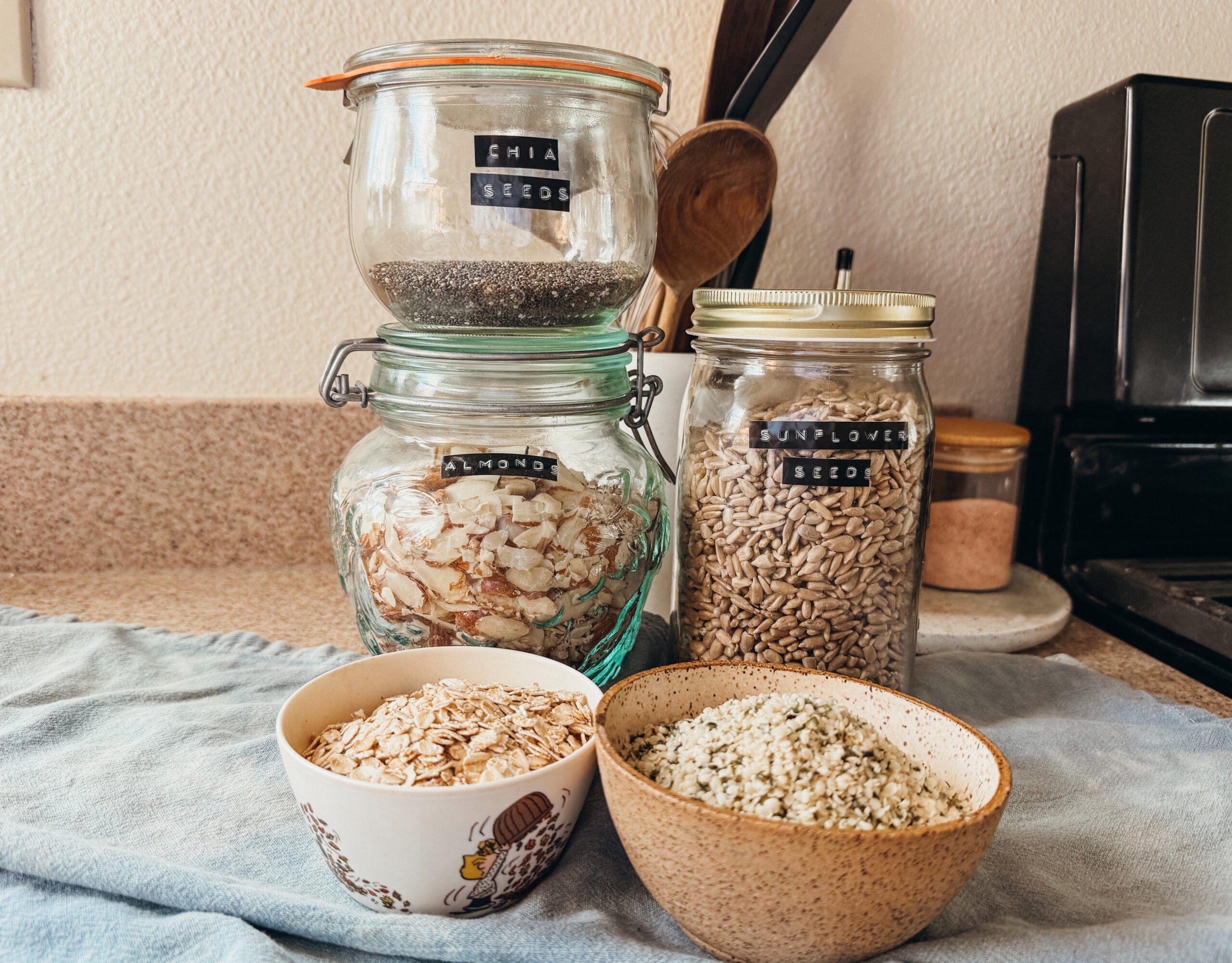 Jars of ingredients for hemp protein powder. Two small bowls sit in front and all are on top of a blue tea towel.