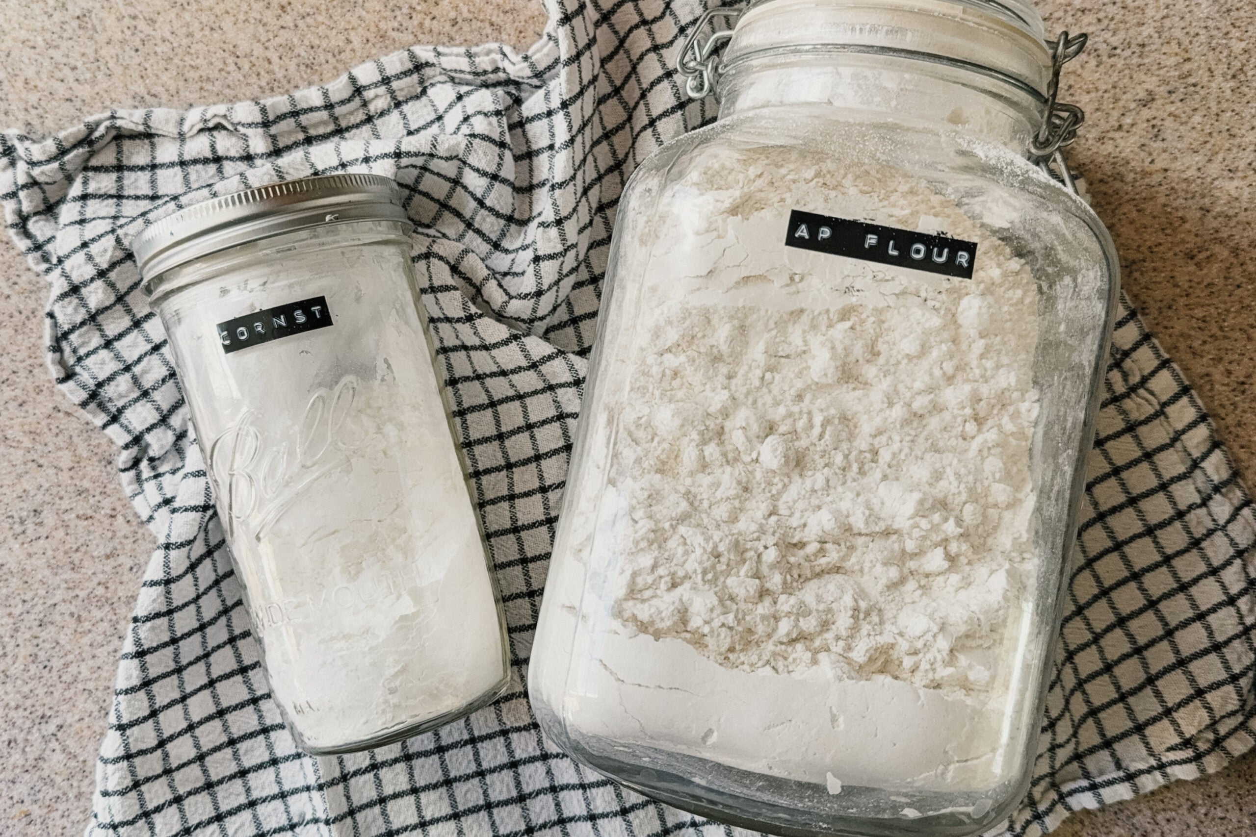 Two large jars of ingredients from left to right: cornstarch, all-purpose flour. A checkered tea towel lays underneath the jars.