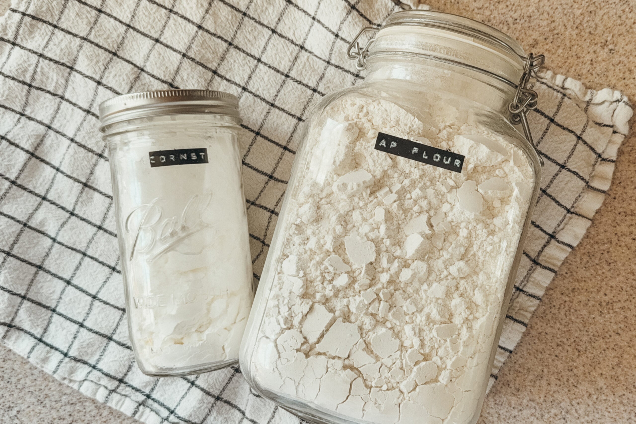Two large jars of ingredients for pastry flour: all-purpose flour on the right, cornstarch on the left. Both jars sit on top of a checkered tea towel.