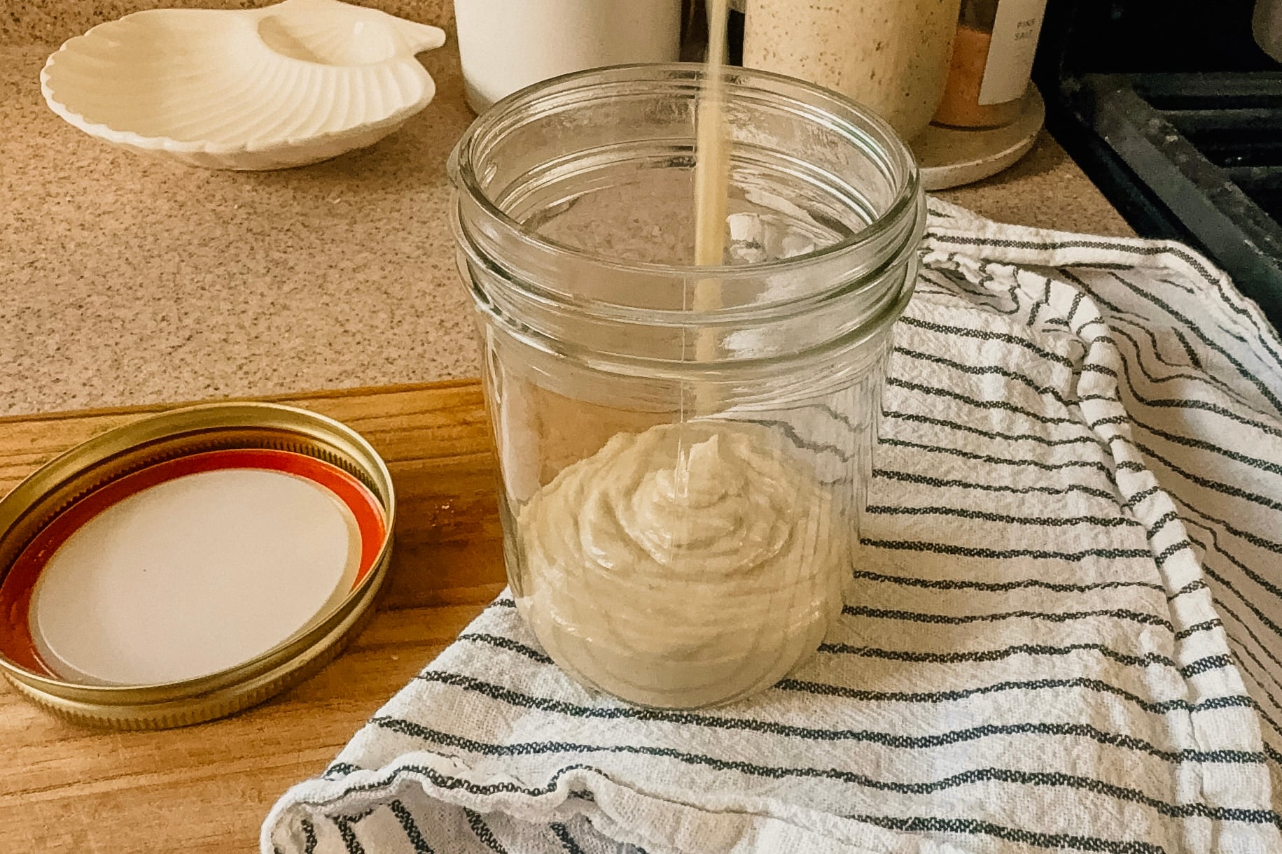 Peanut butter being poured into a glass jar. A striped tea towel is underneath the jar and the lid is to the left side.