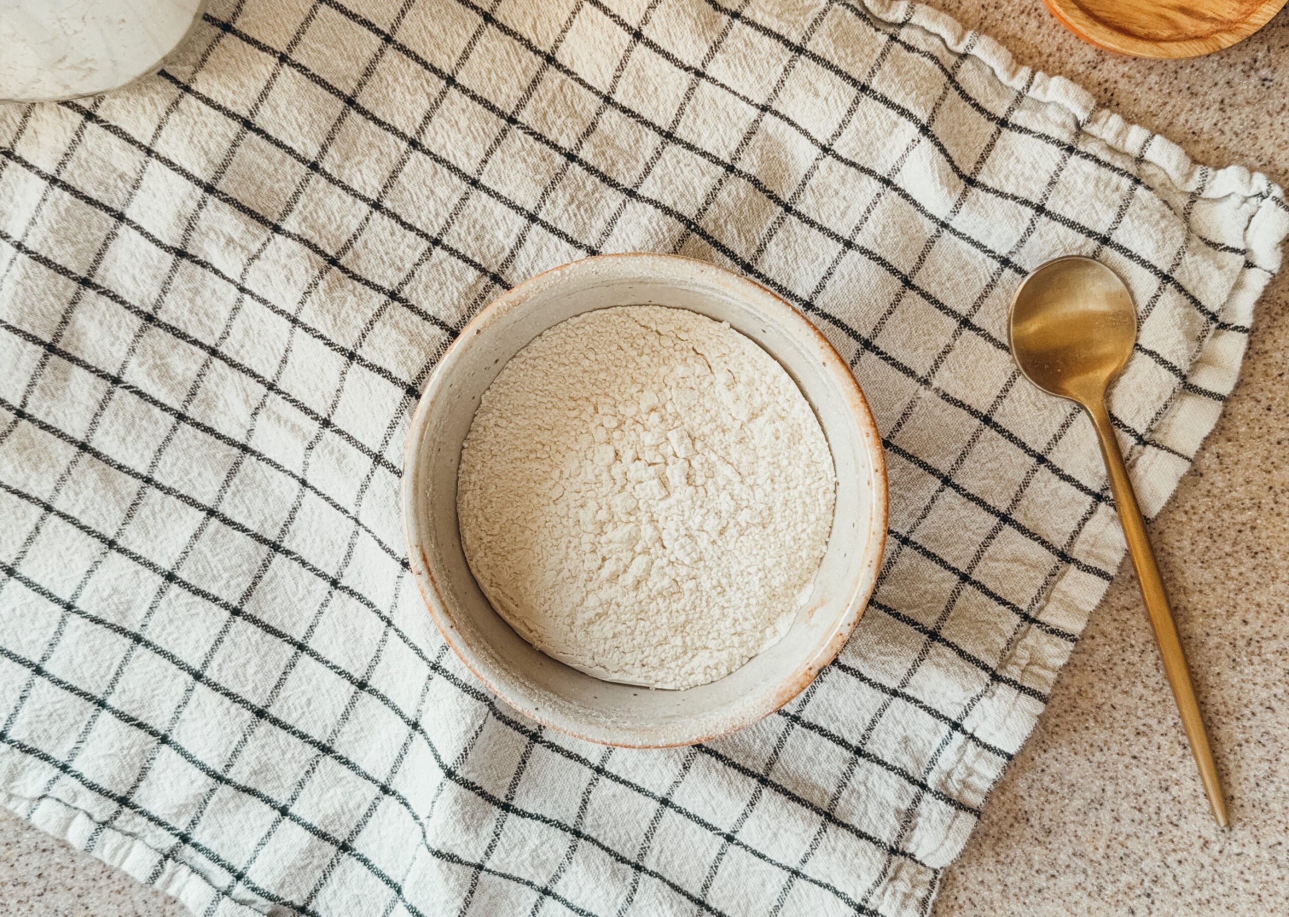 A bowl of flour sitting on top of a checkered tea towel. A gold spoon sits to the right of the bowl.
