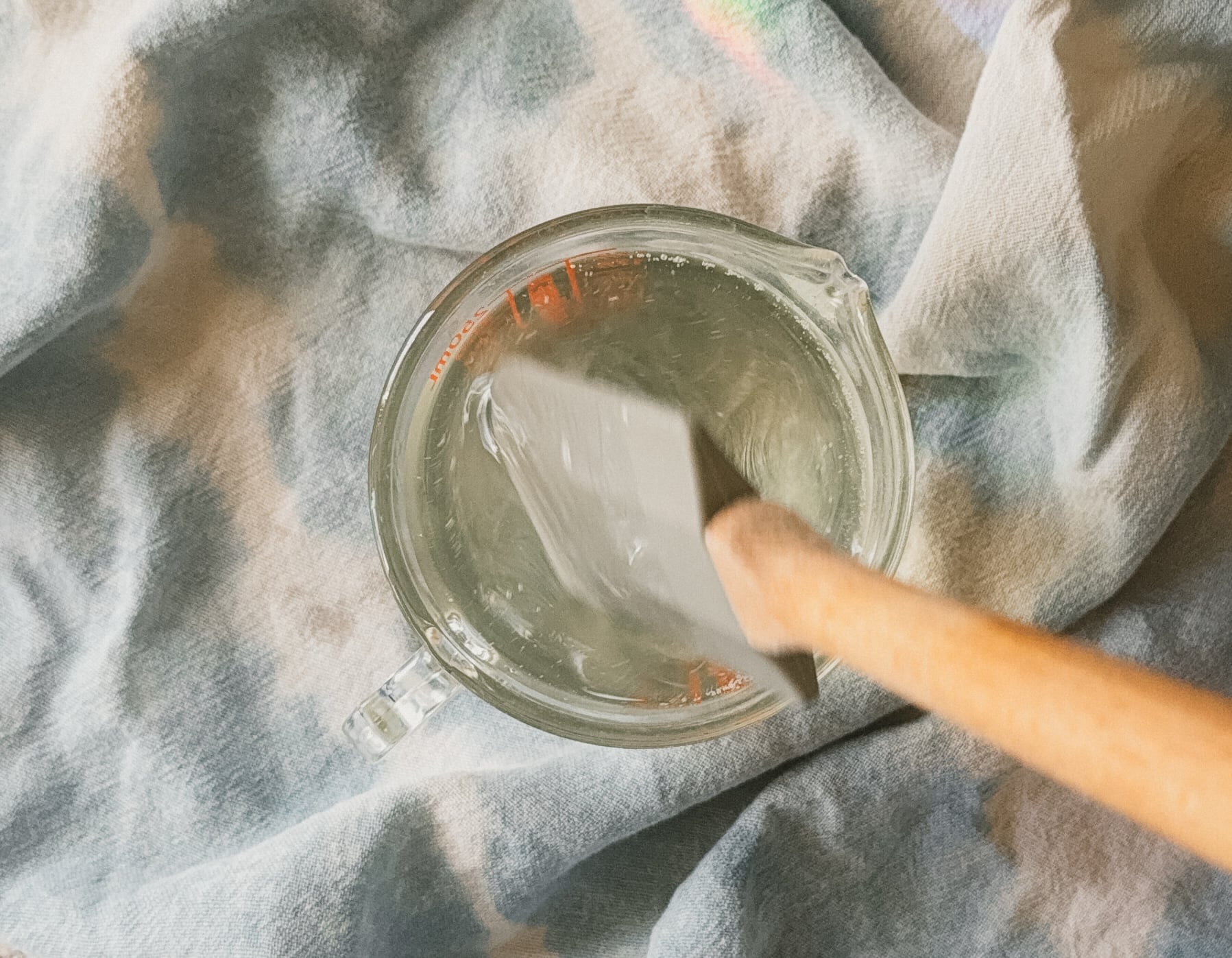 A spatula stirring a glass measuring cup with melted oil. A blue tea towel lays underneath.