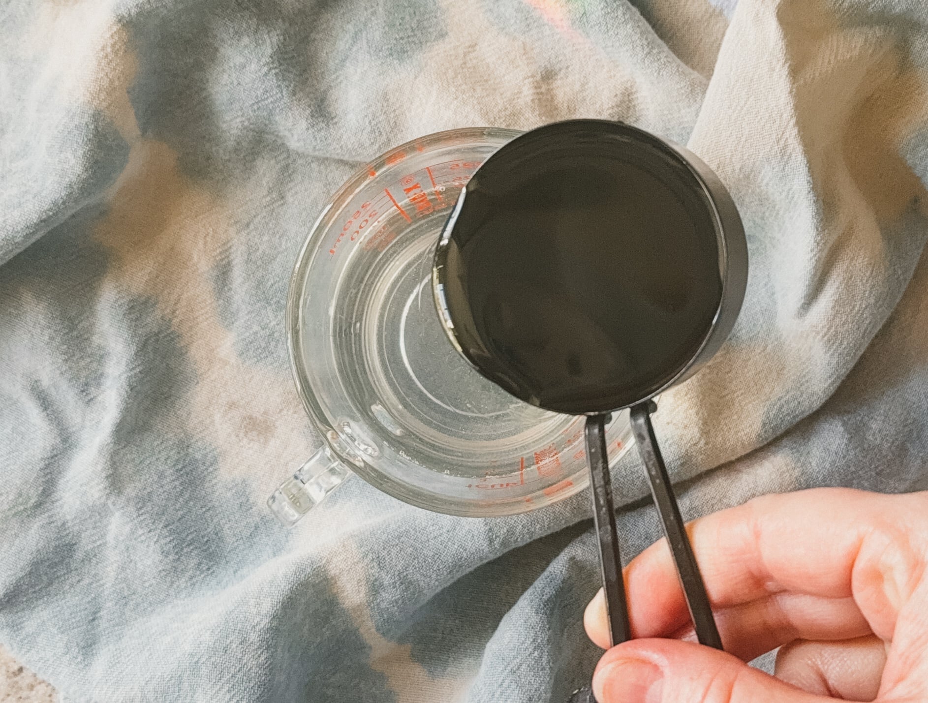 A hand pouring a measuring cup of oil into a glass measuring cup. A blue tea towel lays underneath.