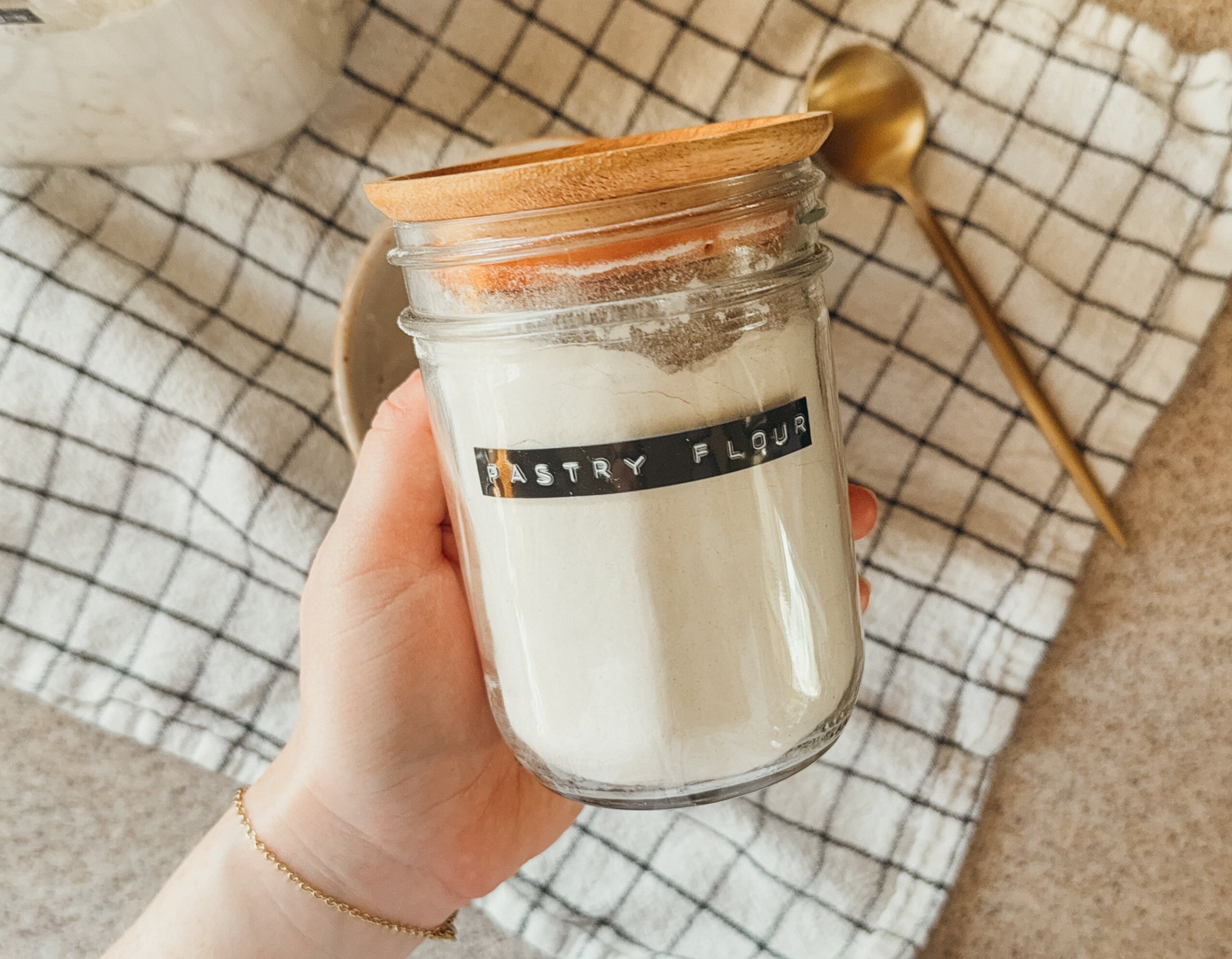 A hand holding a jar of homemade pastry flour above a bowl, gold spoon, and checkered tea towel.