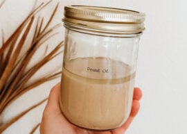 A hand holding a jar of peanut butter & peanut oil in front of a palm frond.