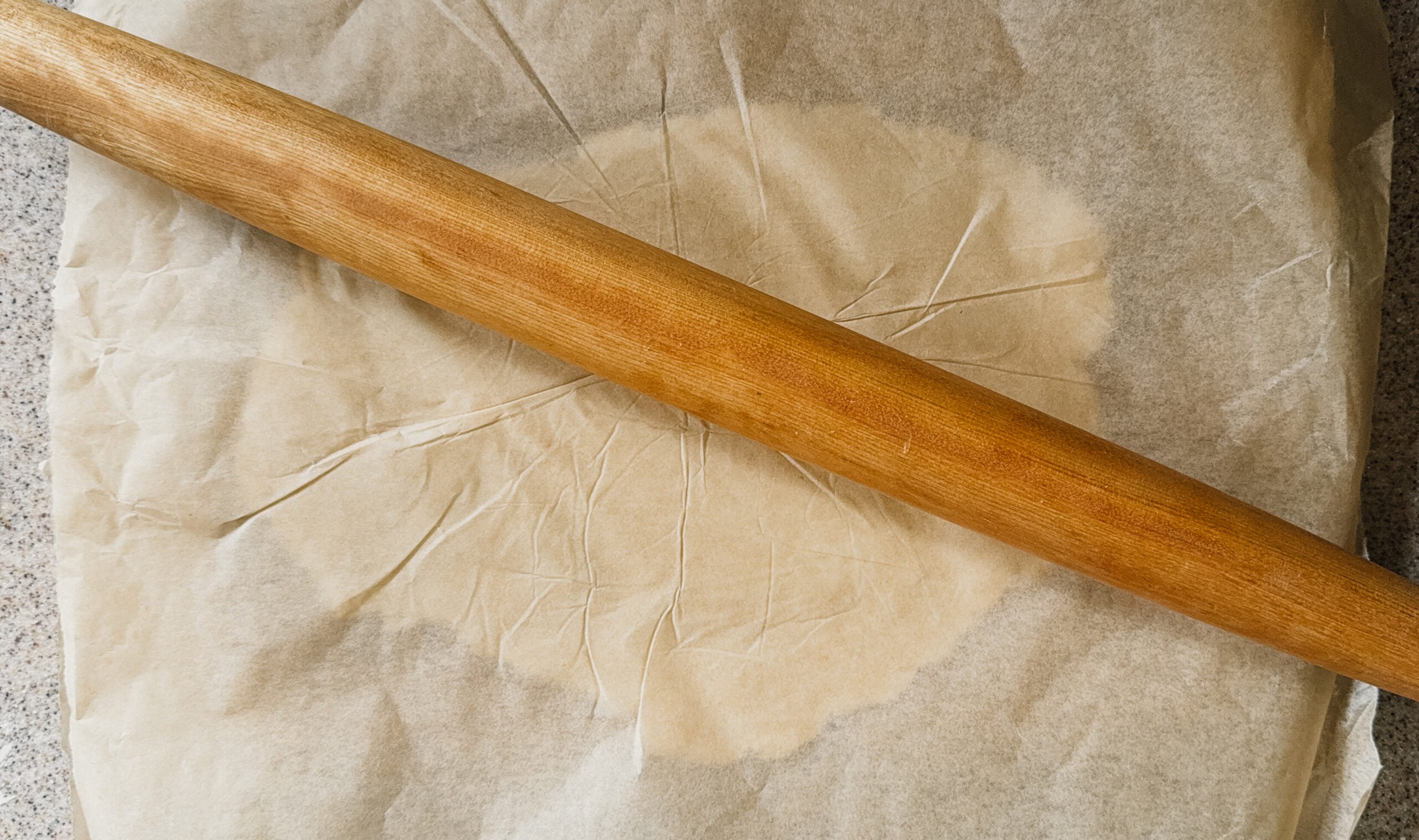 Dough underneath a piece of parchment paper being flattened with a rolling pin.