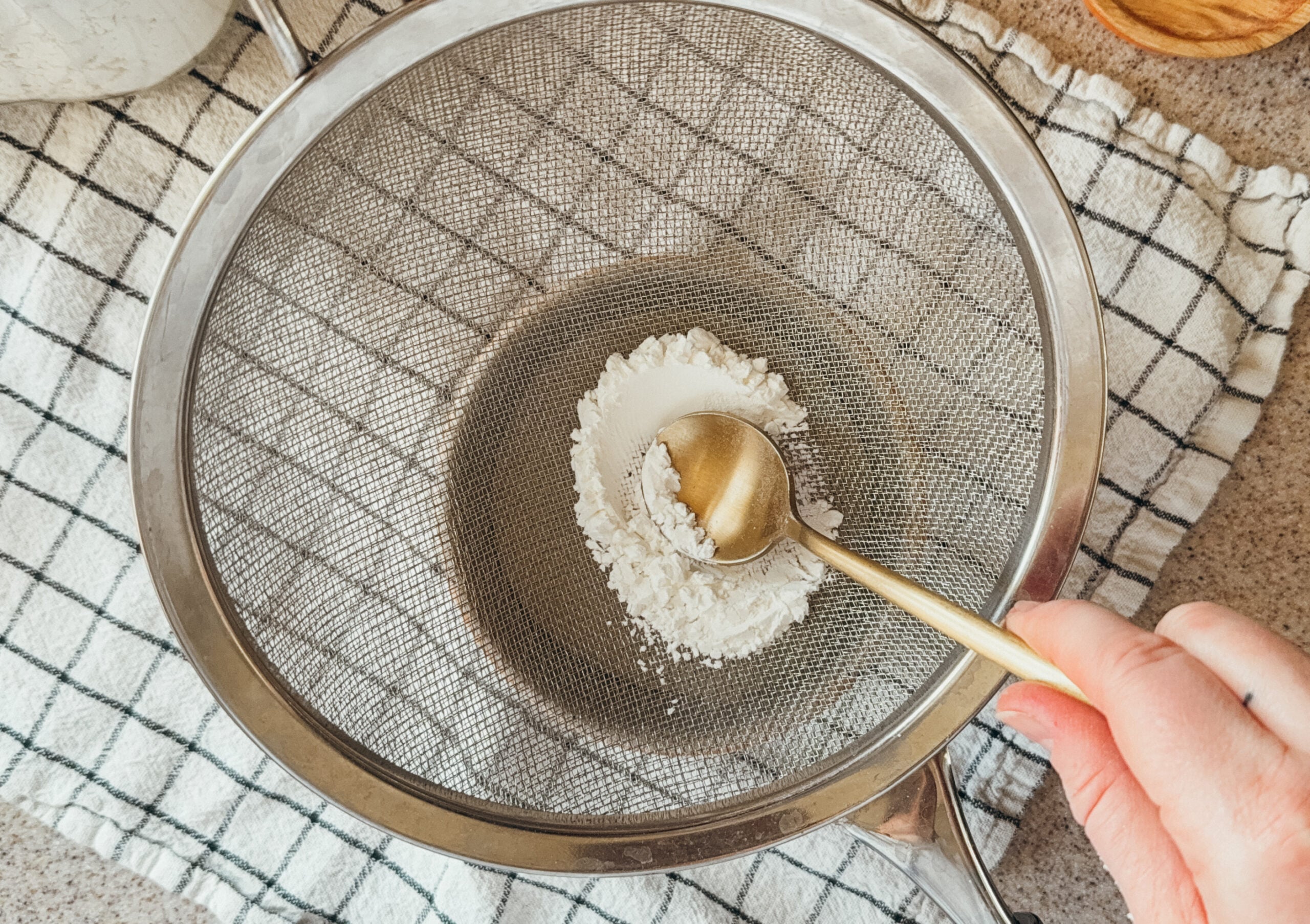 Cornstarch being sifted into a bowl with a gold spoon. A checkered towel sits beneath.