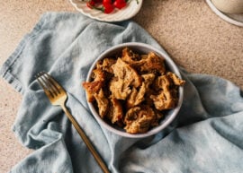 A bowl of cooked vital wheat gluten pieces on top of a blue tea towel. A gold fork sits to the left of the bowl and a plate of small red peppers sits at the top.