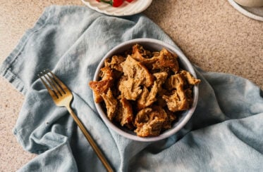 A bowl of cooked vital wheat gluten pieces on top of a blue tea towel. A gold fork sits to the left of the bowl and a plate of small red peppers sits at the top.