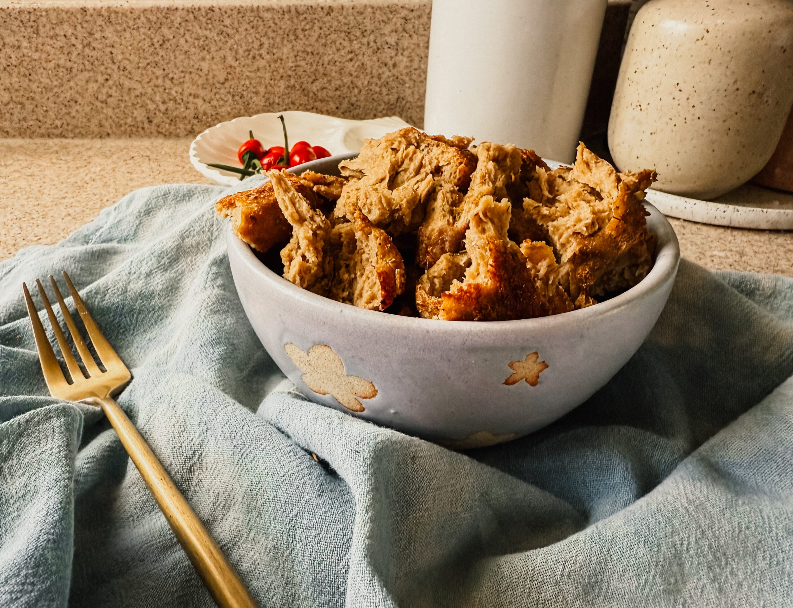 A bowl of cooked vital wheat gluten pieces on top of a blue tea towel. A gold fork sits to the left of the bowl and a plate of small red peppers sits in the back, alongside a crock of utensils.