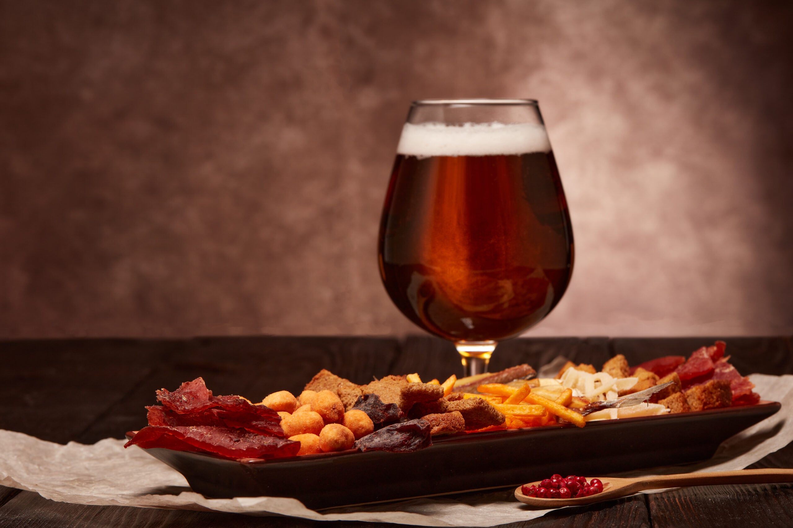 A pint of nut brown ale beer encircled by assorted snacks. 